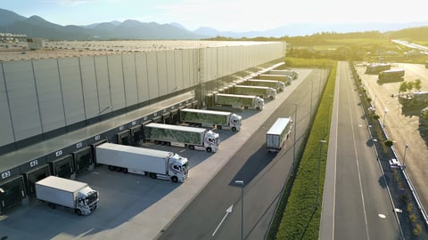 Aerial view of a bustling distribution warehouse with multiple trucks lined up at loading docks with mountain backdrop