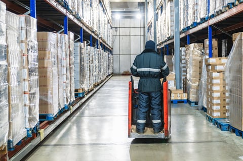 Warehouse worker in protective clothing operating a pallet jack, moving stock within a large, low-temperature cold storage facility