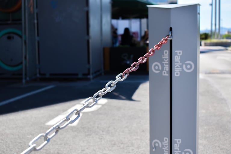 A metal post with red and white striped chain attached, blocking access to a parking area with buildings in background