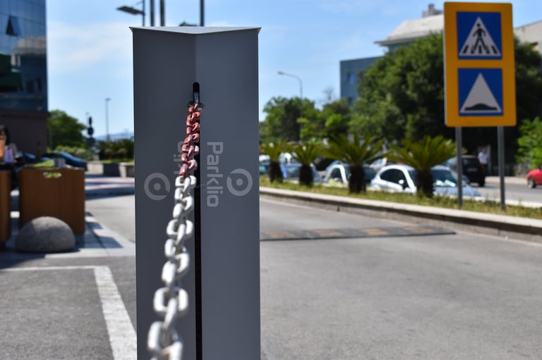 Urban street scene with dark pole featuring vertical chain installation in foreground, blue pedestrian crossing sign visible on yellow post in background